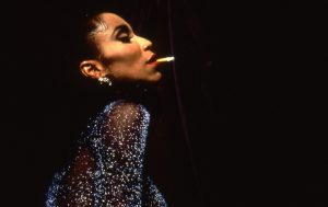 The image displays a drag queen with a cigarette in her mouth wearing a sparkly dress. The background is black. 