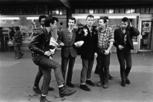 The black and white photo depicts a group of Teddy boys dancing and hanging out outside of a store. There are seven young men in the frame and one is posed with his leg raised.