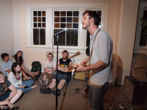 A person singing and playing a guitar at a house show with people sitting on the carpeted floors.