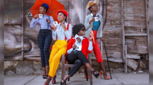 Four Sapeuses pose with mugs, each dressed in bright, stylish clothes, looking off to the side. Two sit, while the other two stand, one holding an umbrella and the other leaning on a cane.