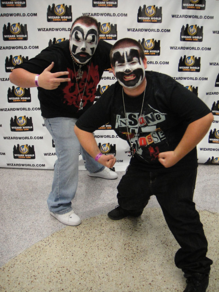 Due giovani Juggalos ad un raduno degli Insane Clown Posse con trucco da clown e magliette ICP in piedi davanti ad uno sfondo.