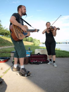 Two people, one Chris Clavin, playing a guitar and violin outside. Chris' guitar reads "Weapon of Mass Destruction"