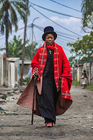 A Sapeuse stands in the middle of a dirt road, close to the camera, wearing a cloak and a top hat, holding a cane. 