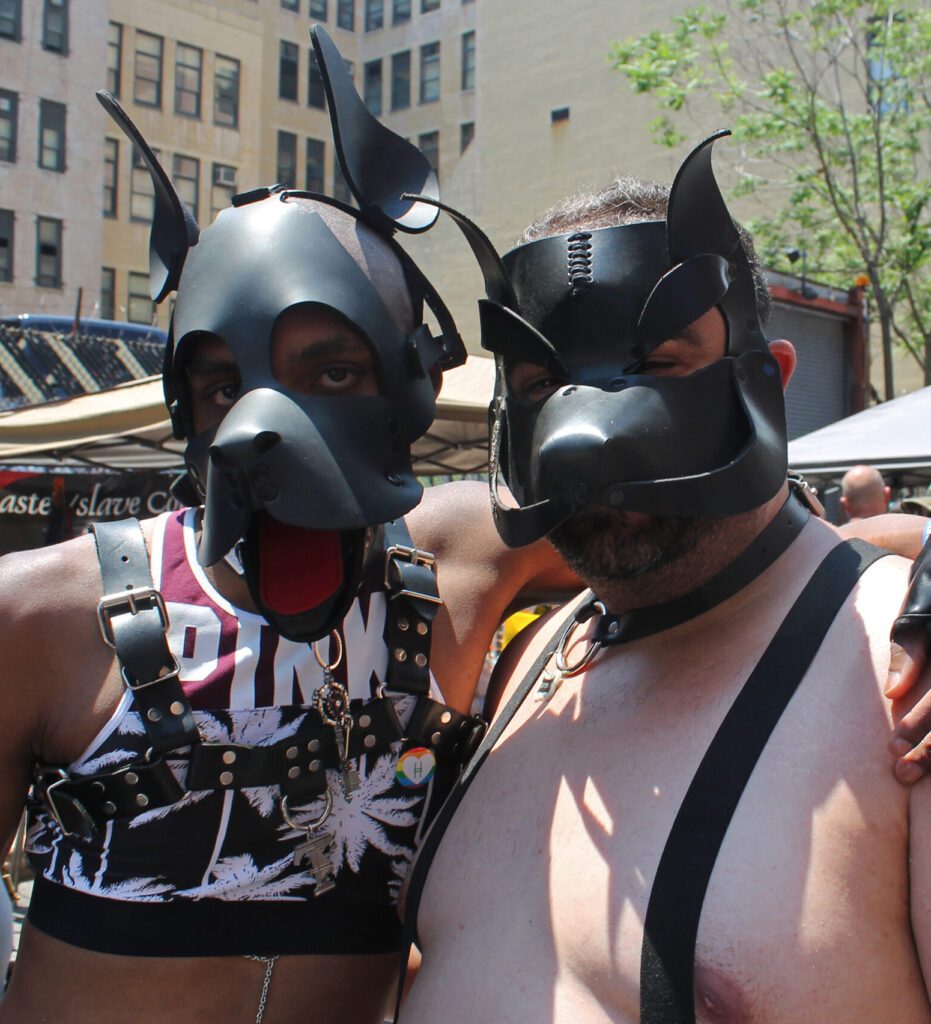 Two people wearing black pup hoods pose at a festival. The person on the left has their arm around the person on the right.