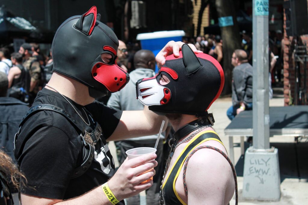 Profile view of two pups in red and black hoods facing each other. The pup on the left is slightly taller and is resting his hand on the other person's head. The pup on the right looks up at the other.