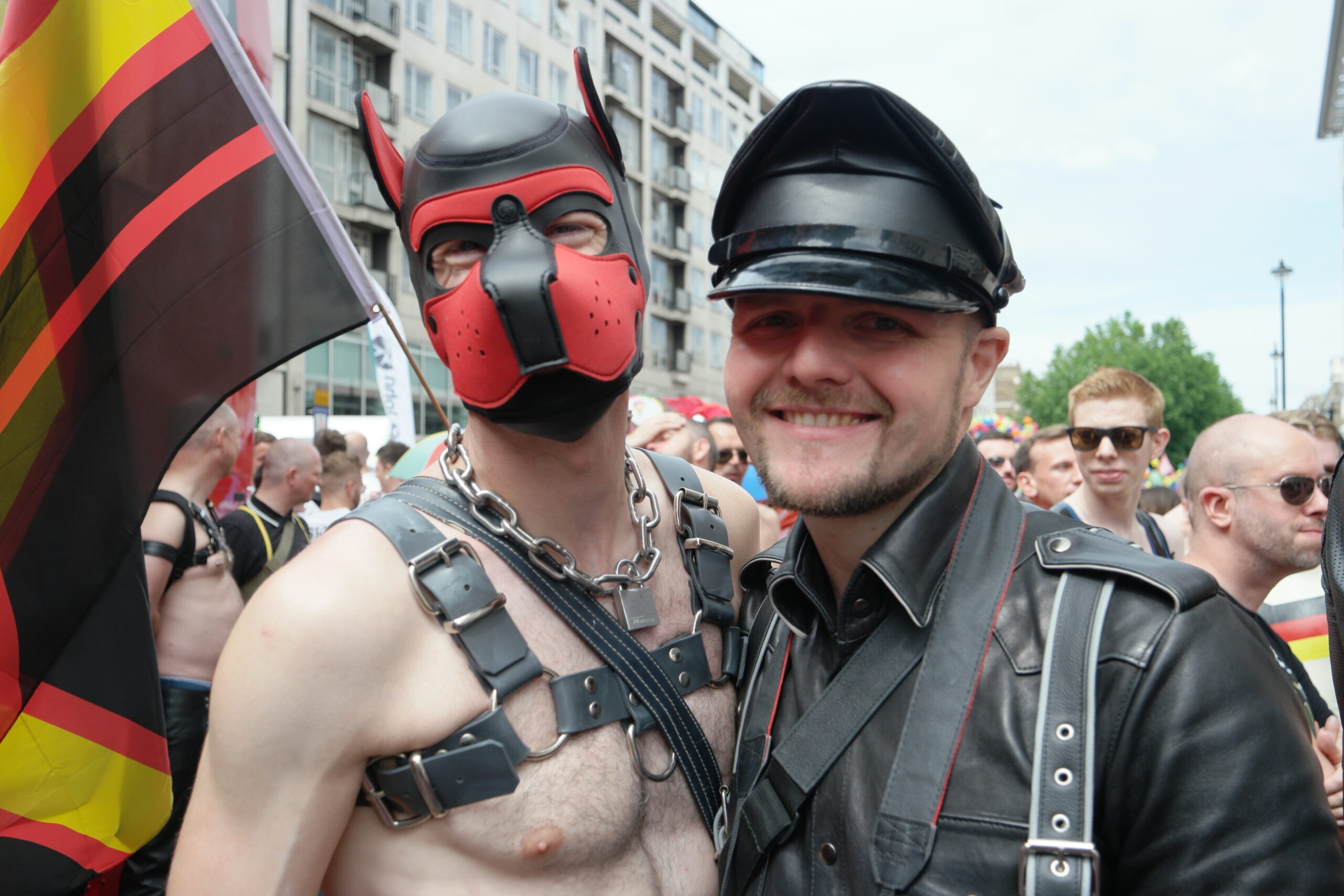 Picture of two people at a festival from the chest up. The man on the left is wearing a black and red pup hood, a chain necklace, and a leather harness. The man on the right wears a black rimmed hat and a black leather shirt. Both men are smiling.