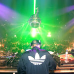 Image showing the back of a DJ in front of a large crowd of people dancing. A disco ball hangs from the ceiling in front of green and purple strobe lights.