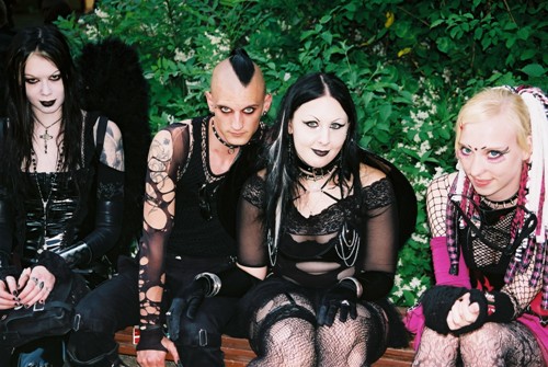 Four people dressed in goth clothing sit outside on a bench.