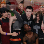 Group of nine young people hugging and making funny faces, dressed in alternative clothing.