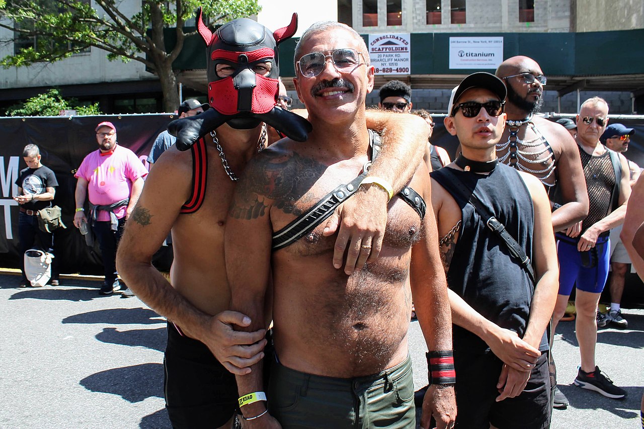 Two people, both shirtless, smile together at a festival. The person on the left is wearing a red and black pup hood and has their arm around the other's shoulder. The person on the right is wearing a leather harness.