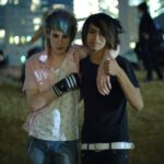 Two young adults smile with their arms around each other. Both have long bangs pushed to the side. The person on the left wears a white tank top, pink shirt, jeans, and fingerless gloves. The person on the right wears a black shirt, black pants, and a white belt.