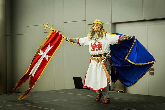 Woman dressed in medieval attire waving a flag with a cross.