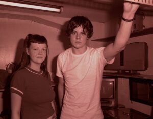 Two young adults stand next to each other. A girl in a red shirt with bangs and a ponytail stands on the left and a boy with a white shirt stands on the right. The picture has a red tint to it.