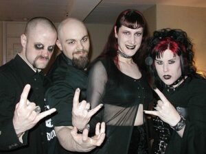 4 goth-dressed people stand in front of the camera a hold up rock-on hand signs. They're all dressed in black, with pale makeup on.
