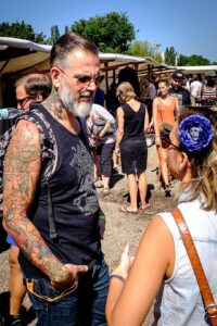 An older man in a black tank top with a full sleeve of tattoos and a gray beard talks to a woman who is nearly off screen.