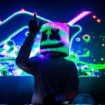 Person behind a DJ station wearing a mask associated with the artist Marshmello. Neon colors and abstract shapes project on the screen in the background.