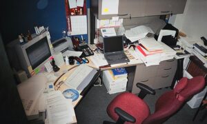 Cluttered office desk with computers, papers, keyboard, pens, pencils, and a chair by the edge