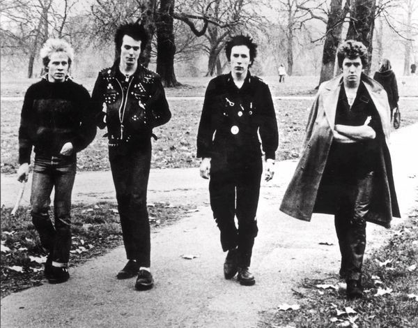 Four men in "punk" clothing walking through the park. They are dressed in all black and have hair that is gelled up.