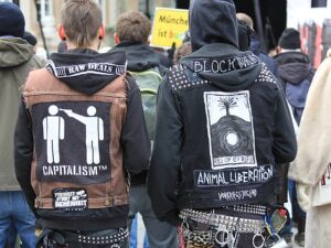 Two people wearing DIY jackets stand with their backs to the camera. The jacket on the left has two stick figures, one raising a gun to the other's head. "Capitalism TM" is printed underneath the figures. The jacket on the right shows text reading "Animal Liberation."