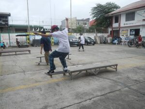A boy in a white shirt, jeans, and a red baseball cap skates on a rail in a parking lot. There are other people around him casually watching.