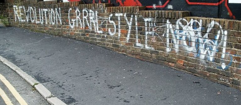 On the side of the road and sidewalk is a brick wall. Spray painted on the wall is "REVOLUTION GRRRL STYLE NOW!"